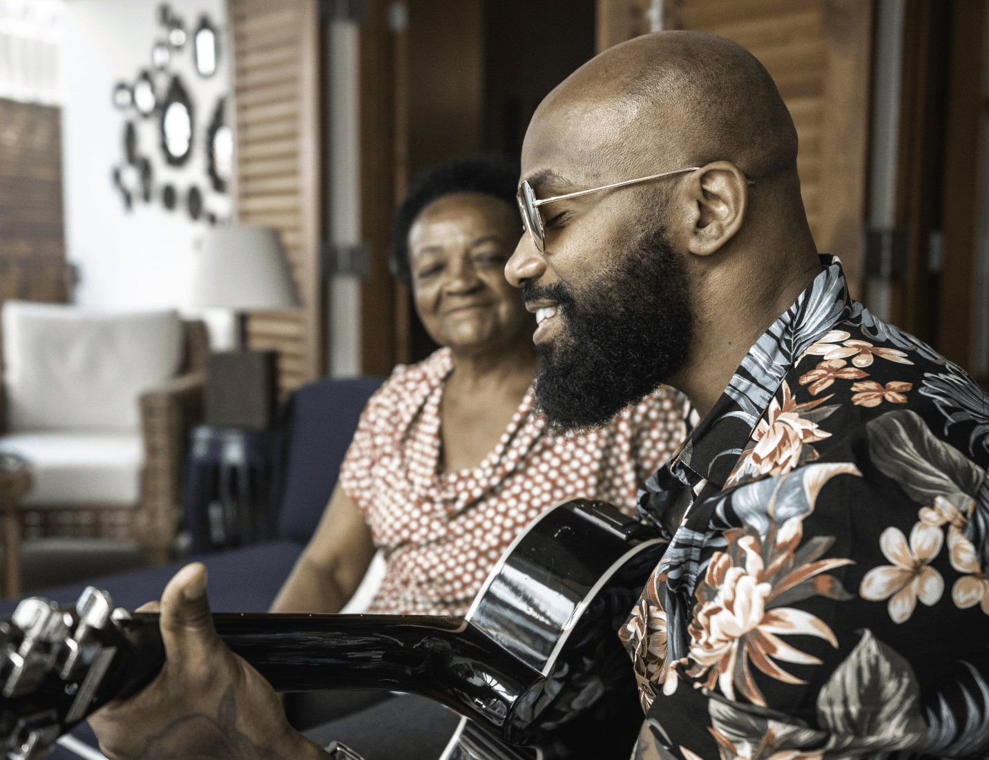 A man plays acoustic guitar in his mom's living room as she proudly watches.