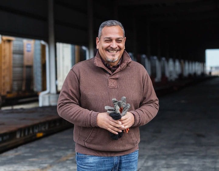 A man holding work gloves smiles as he stands  in front of a truck  loading station.