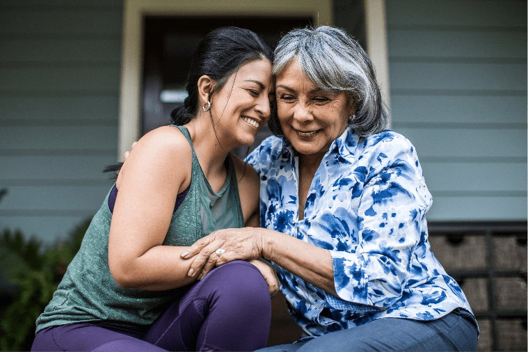 Mother and daughter smile as they embrace while sitting in front of a house.