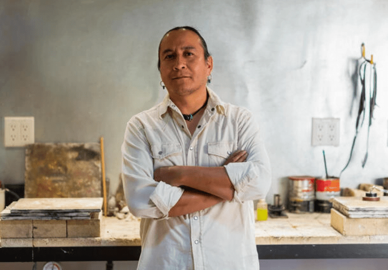 A man, whose face shows confidence, crosses his arms as he leans on his workbench.